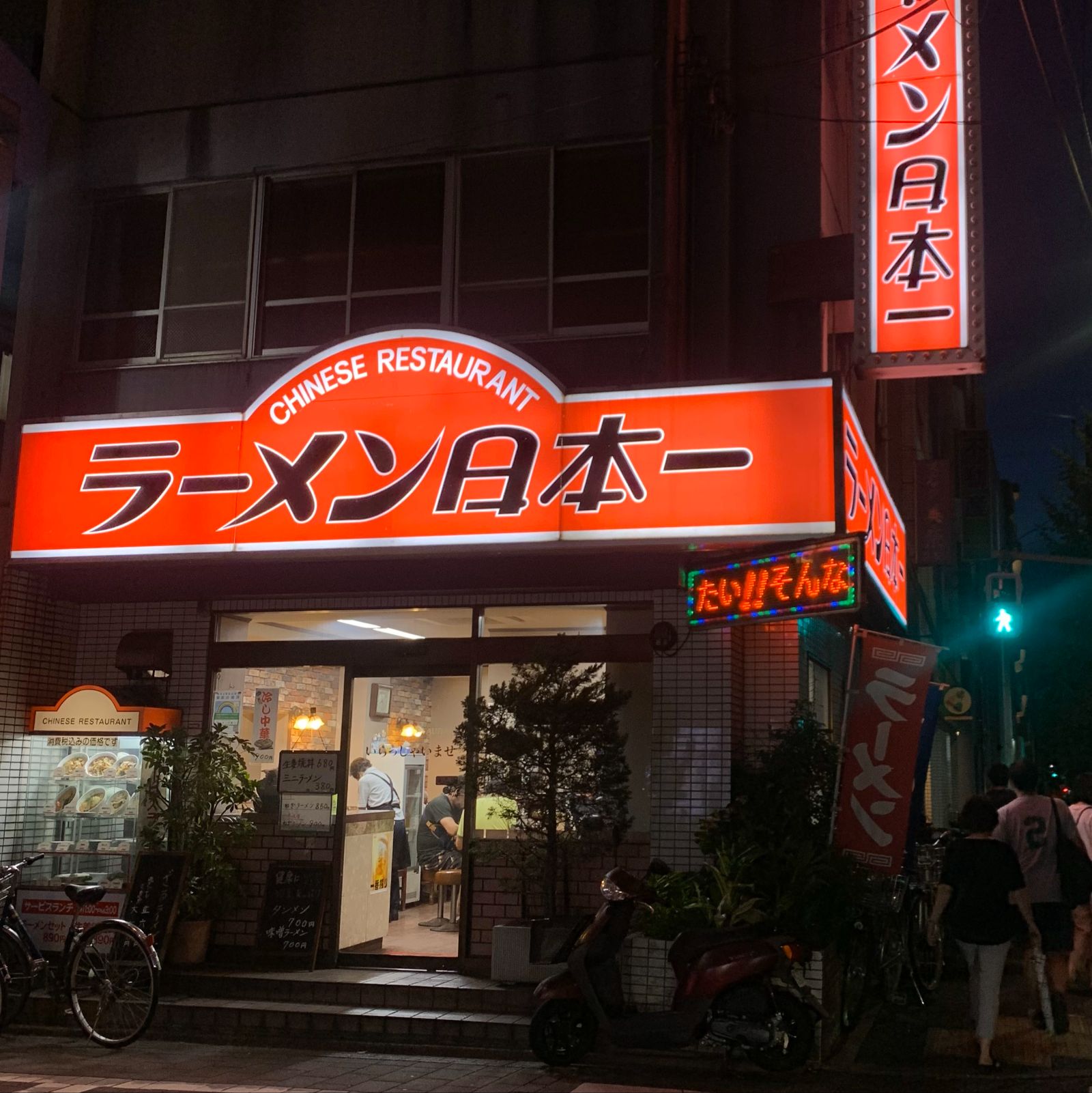 La façade d'un restaurant de ramen, la nuit. Sur la grande enseigne orange, le mot ラーメン est écrit de gauche à droite et son trait est couché. Sur la bannière rouge, à droite, il est écrit de haut en bas et le trait est debout.