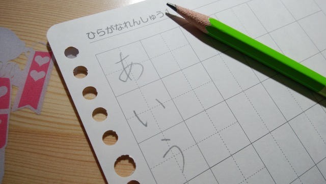 Une page de cahier d'écriture japonais avec あ, い et う tracés au crayon dans les cases, et un crayon vert posé dessus