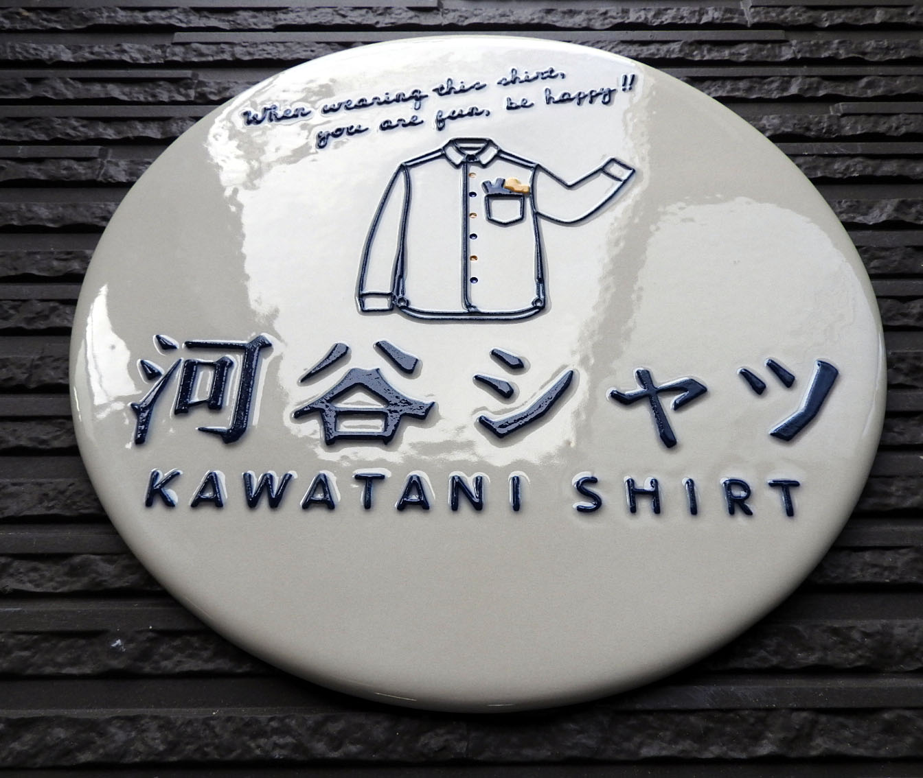 Enseigne ronde en céramique d'un magasin de chemises, avec le mot シャツ écrit en grand sous le dessin d'une chemise