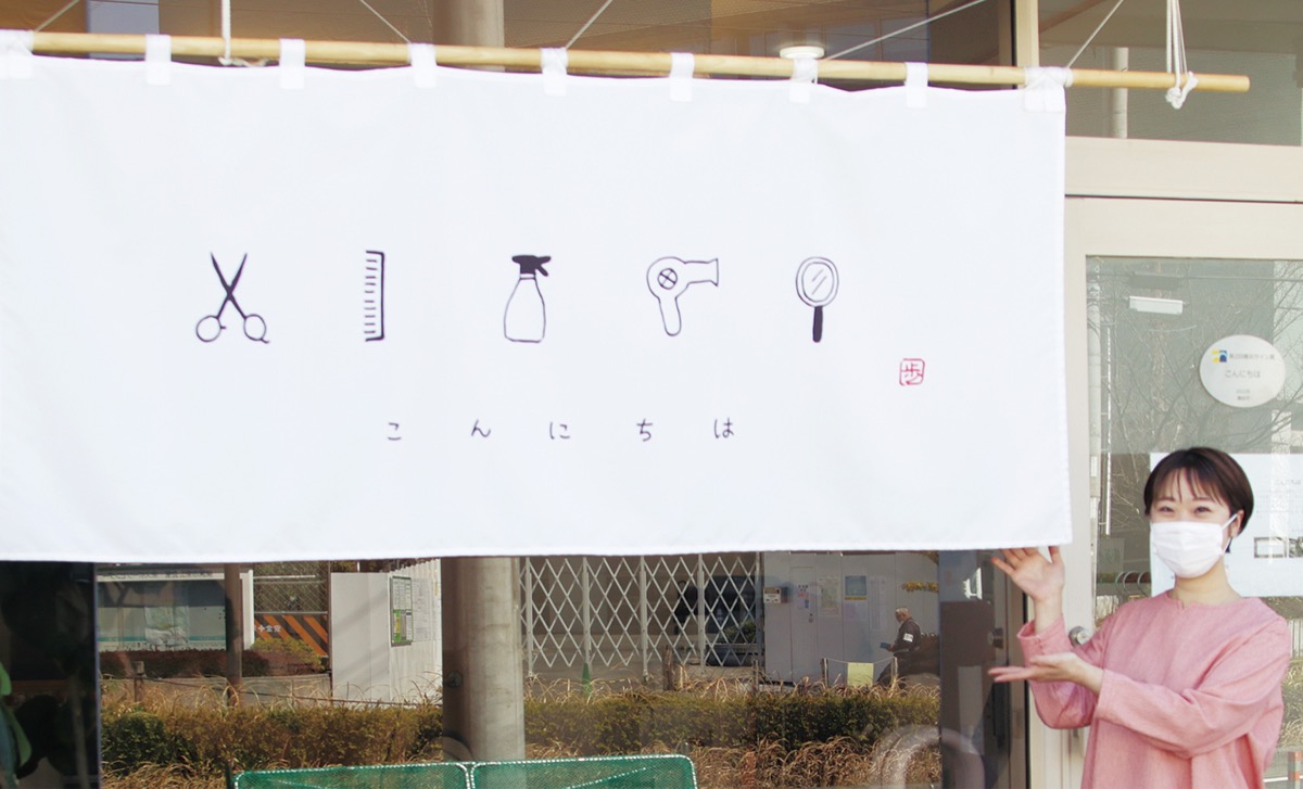 Un noren blanc devant un salon de coiffure japonais, avec des ciseaux, un peigne, un vaporisateur, un sèche-cheveux et un miroir dessinés au-dessus du mot こんにちは écrit en hiragana